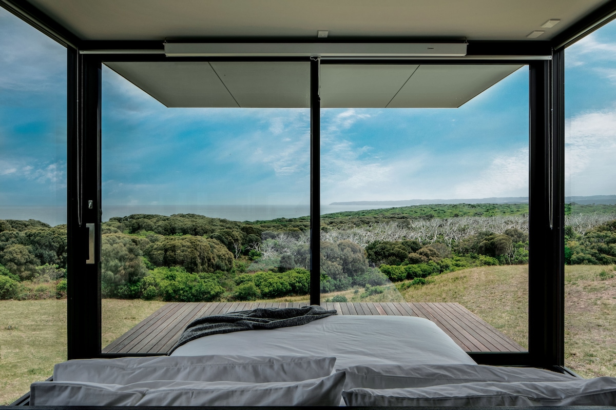 A spacious bed is positioned beside floor-to-ceiling windows, offering expansive views of lush greenery and the coastal landscape. Natural light fills the room, creating a calm atmosphere as the scenery unfolds beyond the glass.