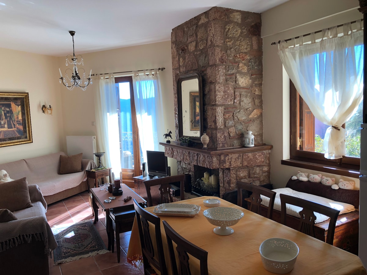 A cozy living area displays natural stone walls, complemented by soft sunlight filtering through sheer curtains. A fireplace and comfortable seating invite relaxation. The dining table is set for gatherings, surrounded by wooden chairs, creating a warm space for shared meals and conversations.
