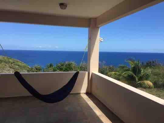 A spacious balcony features a comfortable hammock, framed by white walls. Expansive views of the ocean along with blue sky and greenery are visible in the distance, creating a serene environment for relaxation.