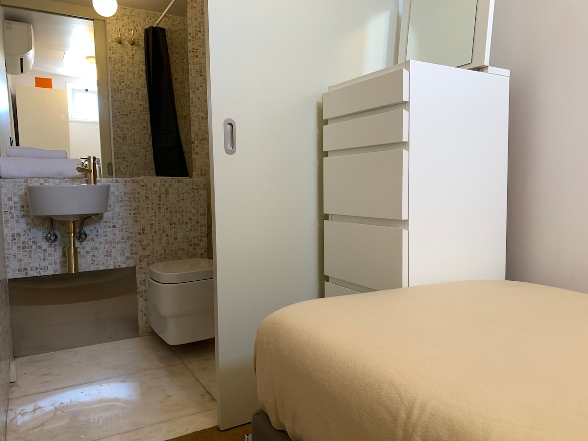 A small suite is showcased, featuring a minimalist design with a light-colored bedspread. A modern dresser stands nearby, and the bathroom is visible through an open door, revealing tiled walls and a sink. Soft lighting creates a calm environment.