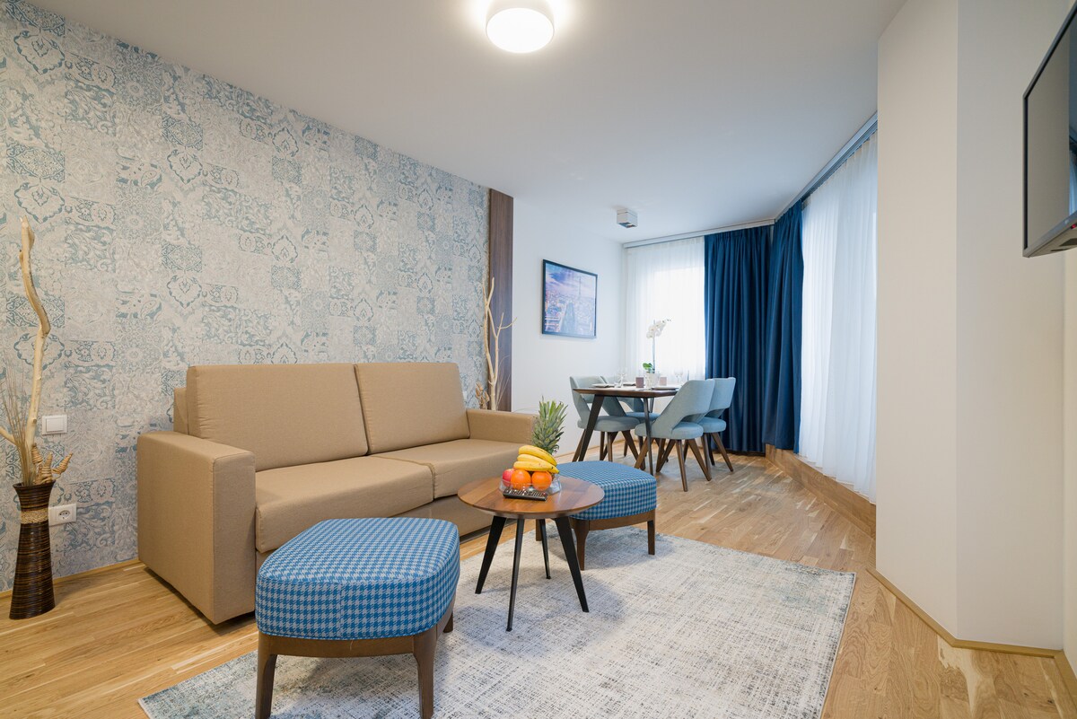 A living area features a comfortable sofa paired with two blue patterned chairs and a round coffee table. Natural light filters through large windows dressed in blue curtains, while a decorative wall with an intricate design adds visual interest.