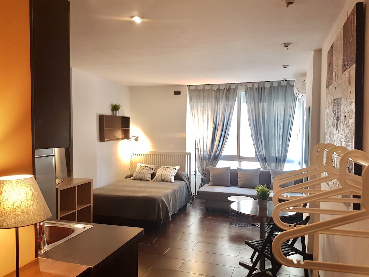 A compact studio features a cozy sleeping area with a spacious bed adorned in neutral tones. A sofa bed and a stylish table are included, along with a light fixture and clothing rack. Large windows allow natural light to fill the room, complemented by soft curtains.