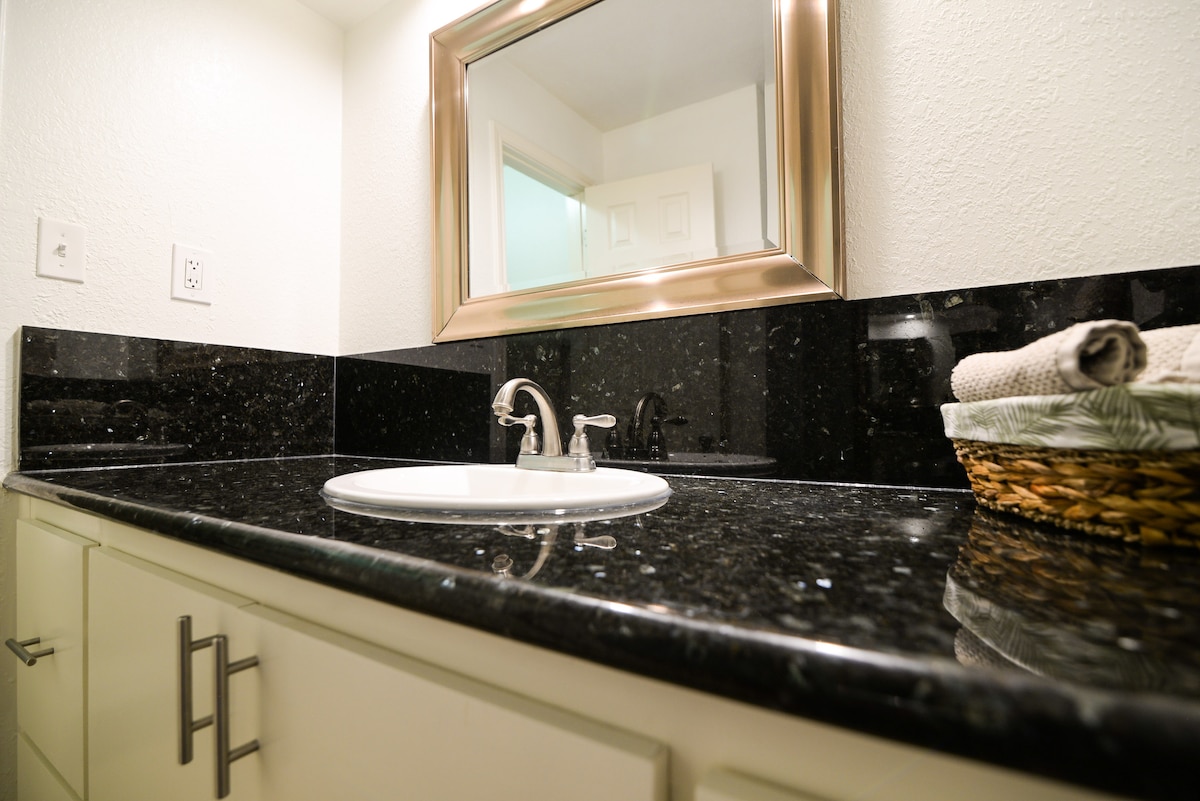 A sleek bathroom countertop features dark granite with shimmering flecks, complemented by a modern mirror framed in bronze. A white sink is centered, while a woven basket holds neatly rolled towels, enhancing the functional and stylish design of the space.