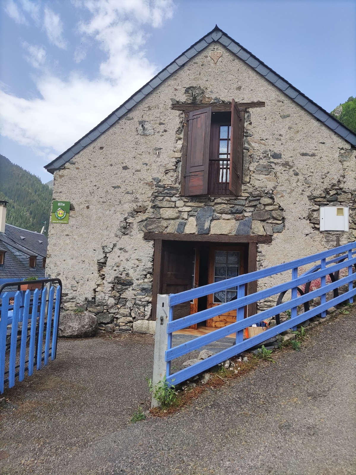 An exterior view of the stone house features a distinct triangular roof and wooden shutters. A blue railing surrounds the entrance, where a wooden door is partially open, revealing a glimpse of the interior. The surrounding landscape includes mountains in the background.