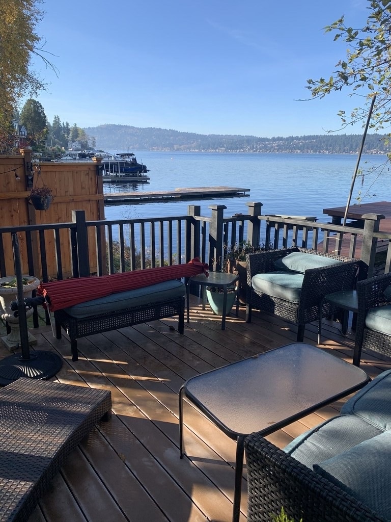 A spacious patio area features comfortable outdoor seating, including cushioned chairs and a low table. The view extends to the tranquil waters of the lake, with a wooden dock visible in the distance. The surrounding landscape includes trees and a wooden fence.