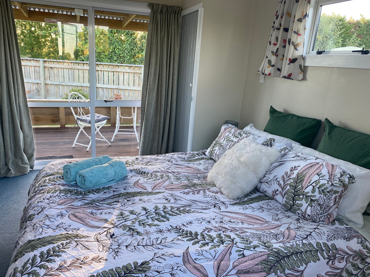 A spacious bed with decorative pillows and a soft throw is positioned near sliding glass doors. Natural light fills the area, revealing a private deck outside. The curtains are elegantly draped, complementing the serene cottage decor.