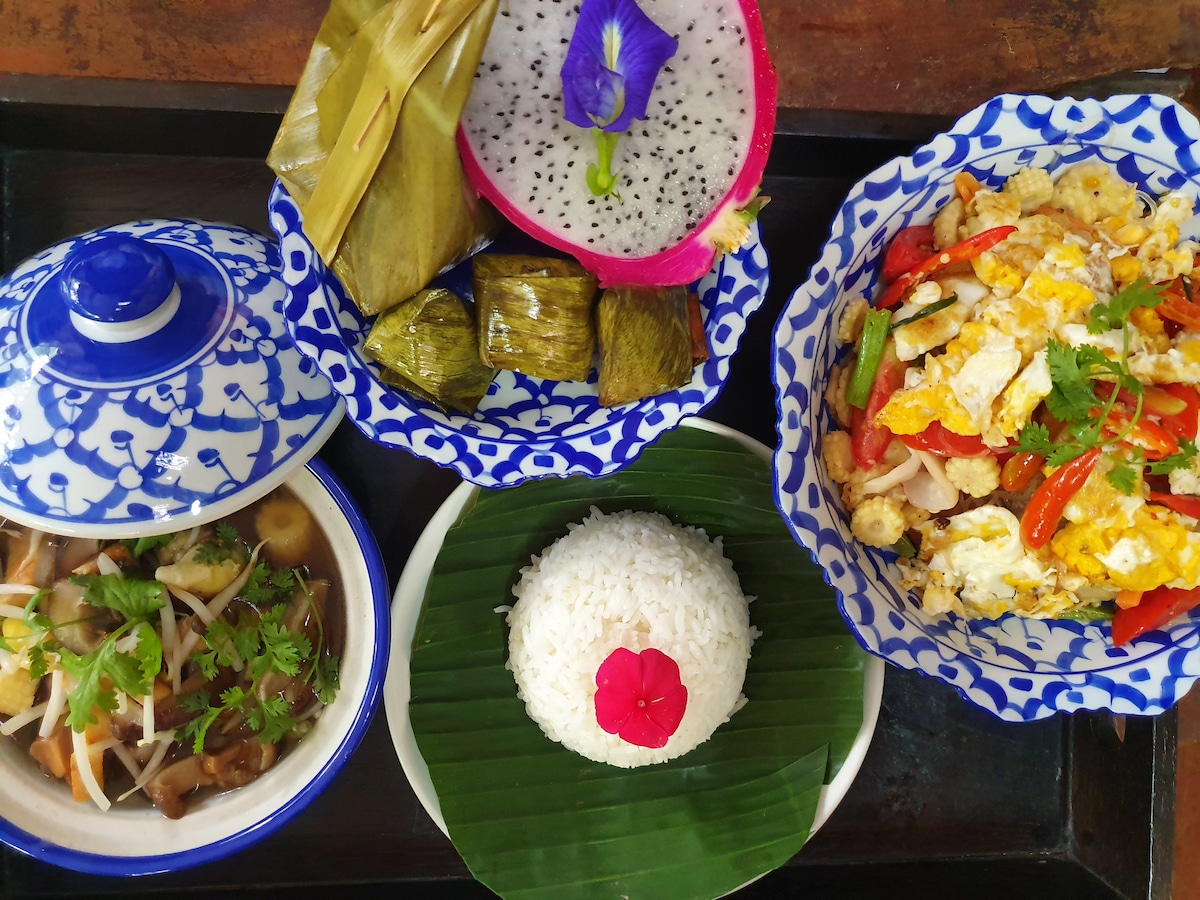 A vibrant meal presentation features multiple dishes arranged on a decorative tray. Blue and white patterned bowls contain stir-fried vegetables and an egg dish. A fragrant white rice mound with a flower garnish is placed on a green banana leaf, alongside traditional wrapped snacks and a colorful fruit piece.