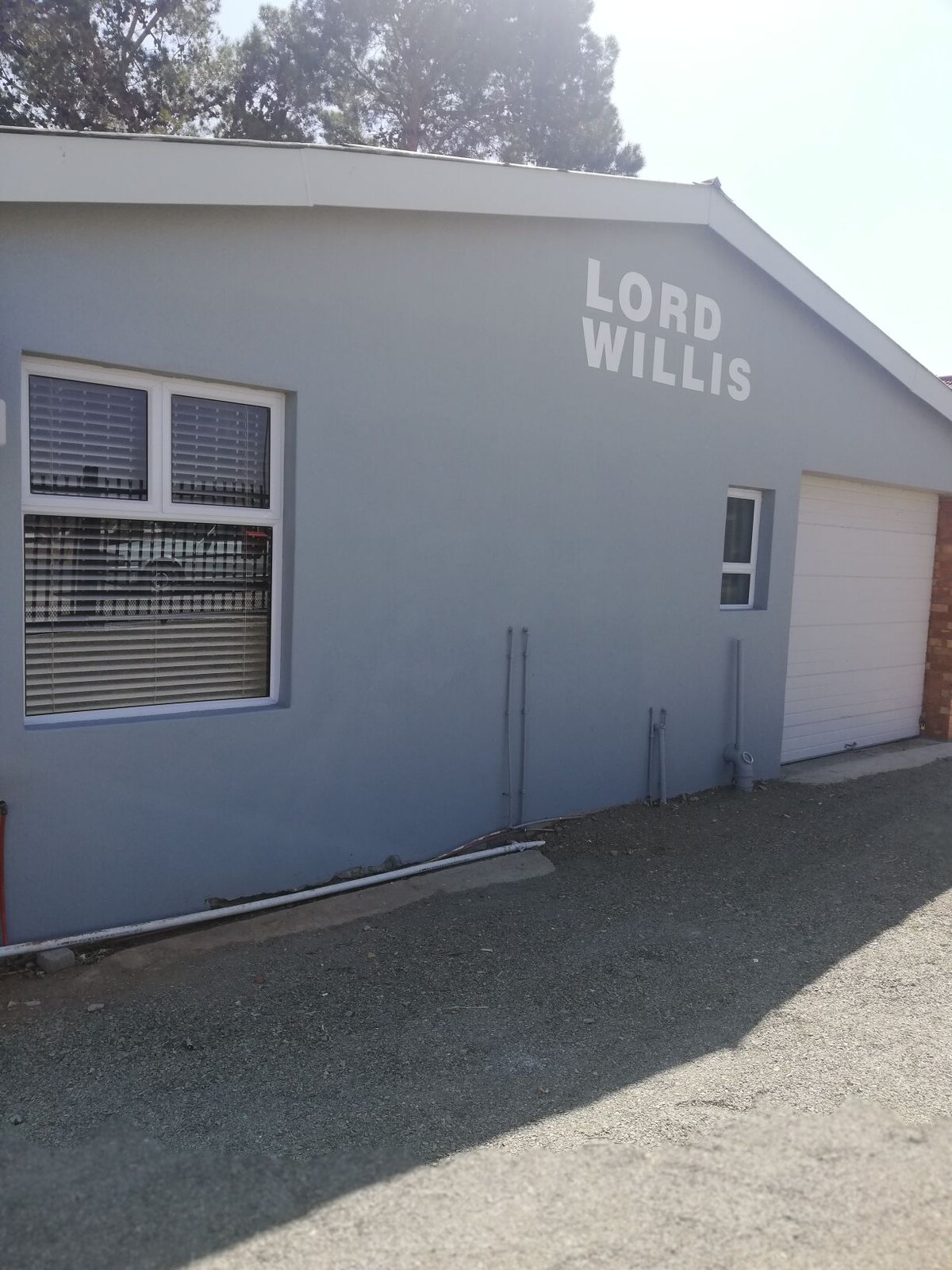 The exterior of the building features a light blue facade with the name 'Lord Willis' displayed prominently above a garage door. Windows with horizontal blinds allow natural light in, while the surrounding area is covered in gravel.