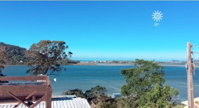 A serene view of the lagoon is presented, showcasing calm waters surrounded by lush greenery. The horizon features distant coastal areas and a clear blue sky, enhancing the tranquil atmosphere. A sun graphic indicates clear weather, contributing to a sense of peace.