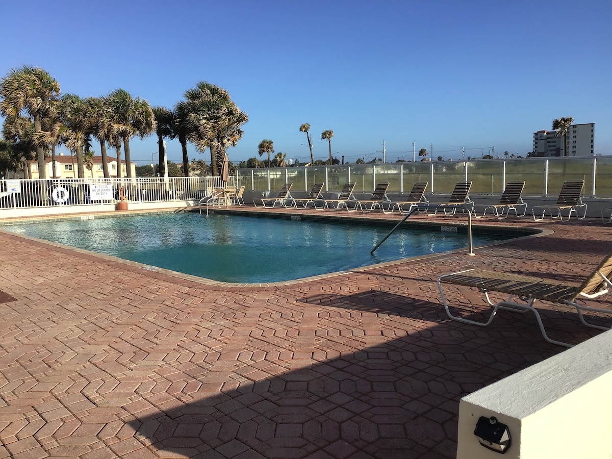 A clear view of the heated swimming pool is presented, surrounded by palm trees and a spacious deck area. Several lounge chairs are arranged along the edge of the pool, with the horizon of the coast visible in the background under a bright blue sky.