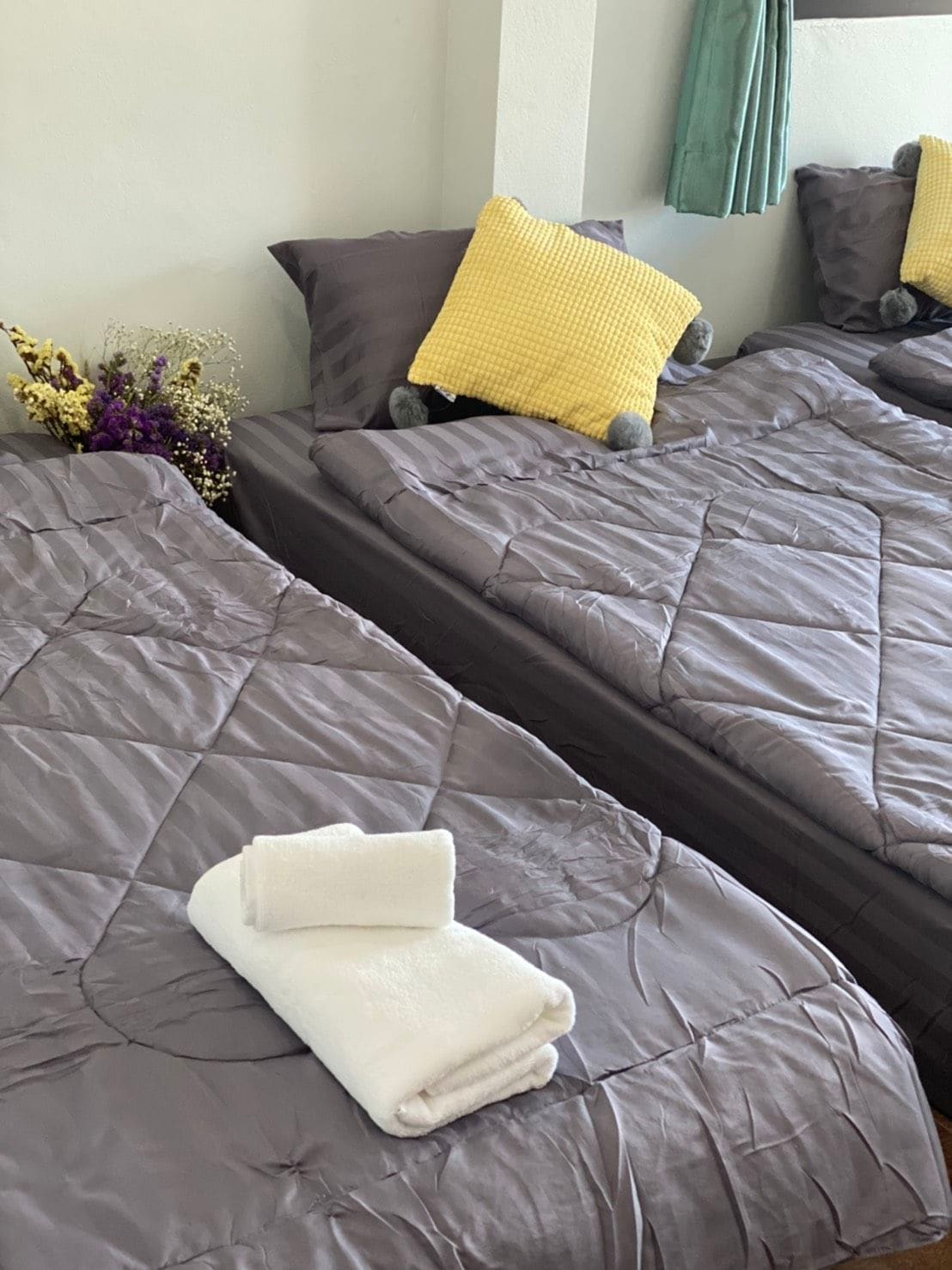 Two spacious beds are arranged in a well-lit room, each featuring soft gray bedding. A yellow checkered pillow adds a pop of color. Neat white towels are placed on one of the beds, while decorative flowers are positioned nearby, enhancing the cozy setting.