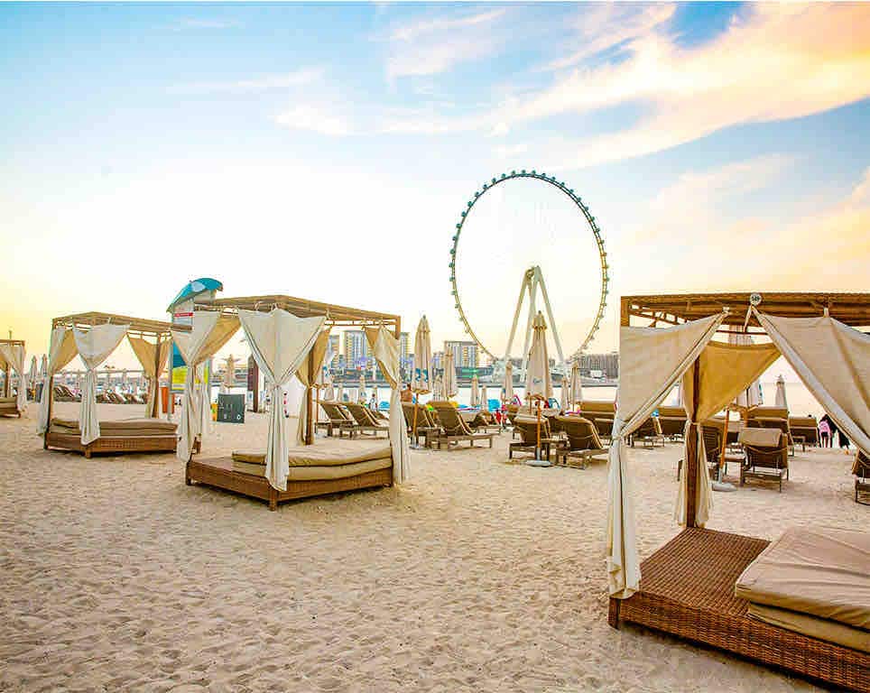 Lounge areas are arranged on a sandy beach, featuring canopied daybeds set on wooden platforms. In the background, a large ferris wheel is visible, along with a skyline of modern buildings. Warm hues from a sunset illuminate the scene, enhancing the tranquil atmosphere.