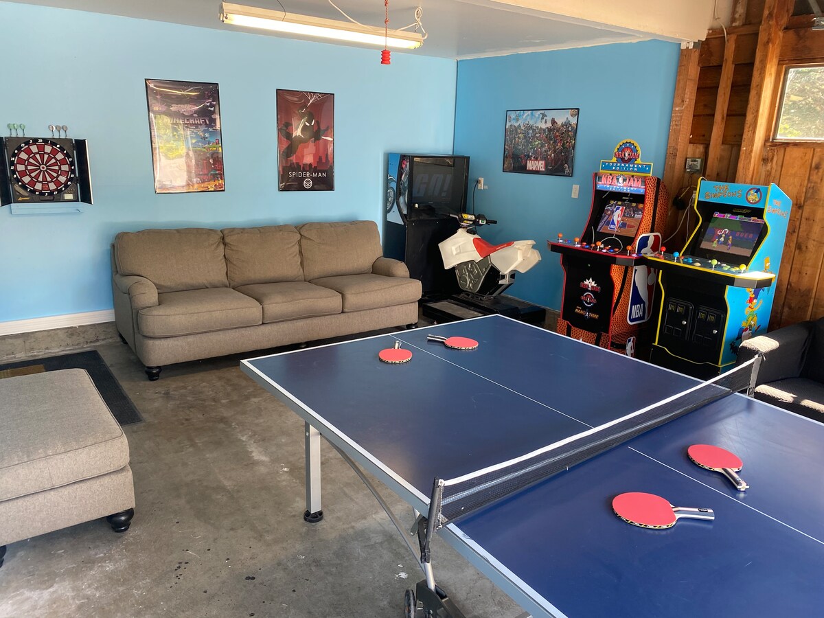 A game room is featured with a ping pong table at its center, complemented by a comfortable sofa and seating area. Colorful arcade machines and a dartboard are positioned against the wall, providing a recreational space for guests. Natural light fills the area through the garage door.