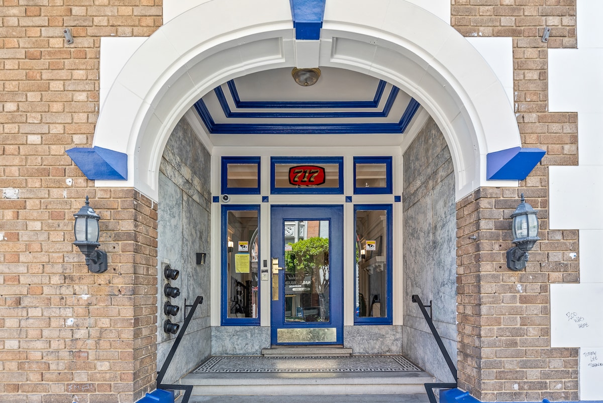 An entrance features an archway framed by blue and white detailing, with brick walls and large glass doors. Two wall-mounted lamps illuminate the entryway, while steps lead up to the door. A decorative mat is placed at the entrance.