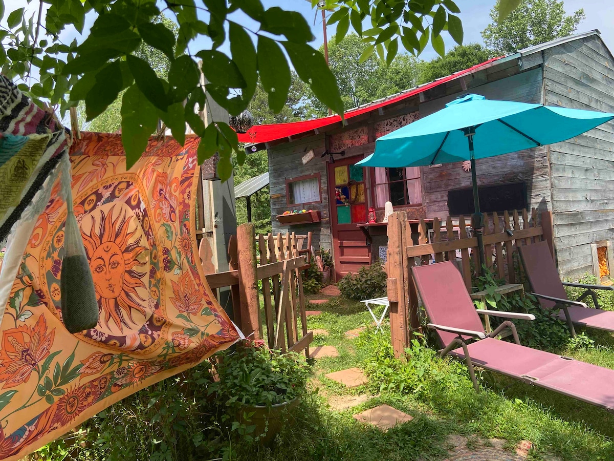 A vibrant courtyard features a cozy guest cottage with a red roof and colorful outdoor accents. Lounge chairs sit among green foliage, while a patterned tapestry hangs nearby. Bright umbrellas provide shade, creating an inviting outdoor space surrounded by a rustic wooden fence.