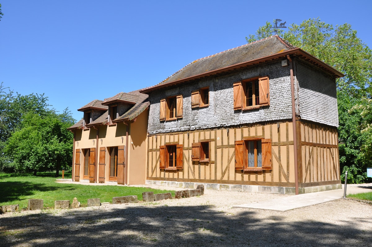 A two-story building features a mix of wooden beams and rustic stone. Window shutters in rich brown are visible, adding character. The structure is surrounded by green trees and a well-maintained lawn, providing a serene outdoor setting.