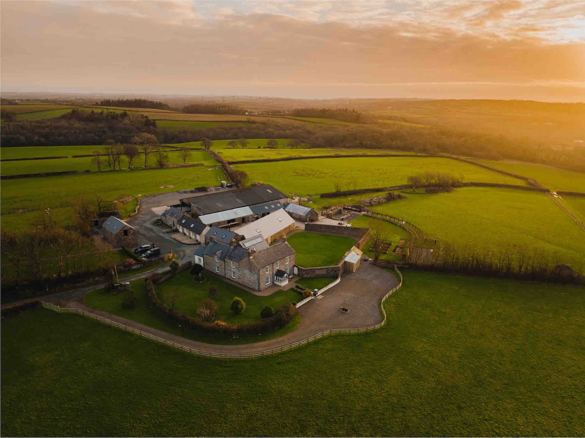 An aerial view captures the expansive countryside surrounding the stable cottage. Vibrant green fields extend into the distance, with a mixture of stone buildings and farm structures neatly arranged in a circular layout, all under a soft evening sky.