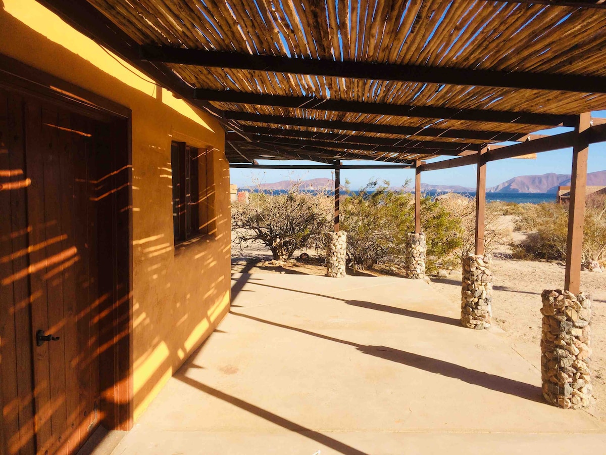 A sunlit patio area features a textured concrete floor and stone columns supporting a wooden pergola. The natural light casts soft shadows, while a view of surrounding greenery and distant mountains extends beyond the perimeter of the structure.
