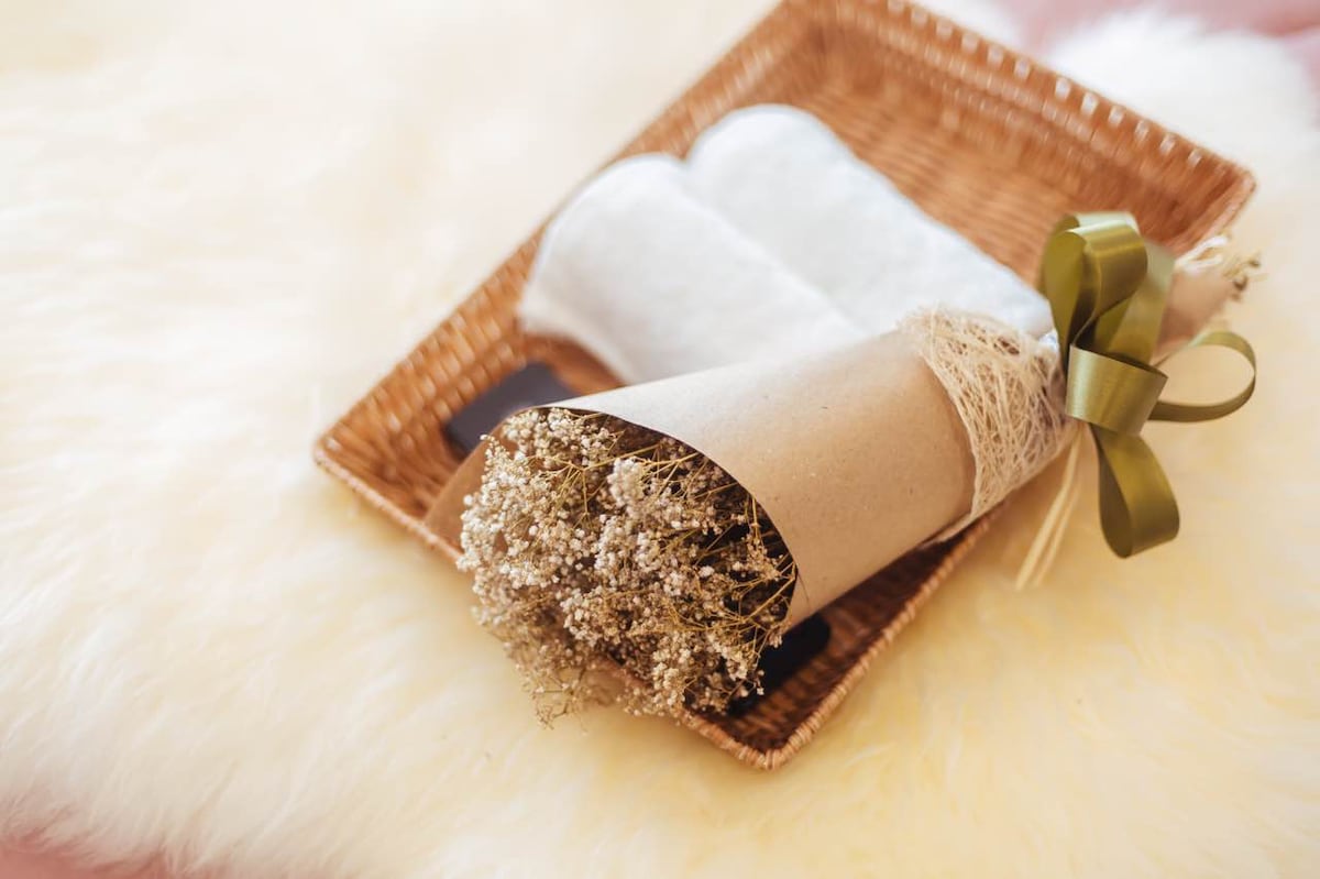 A woven tray holds a bouquet of dried flowers wrapped in paper with a decorative ribbon. Two white towels are neatly placed beside the bouquet, set against a soft, textured surface.
