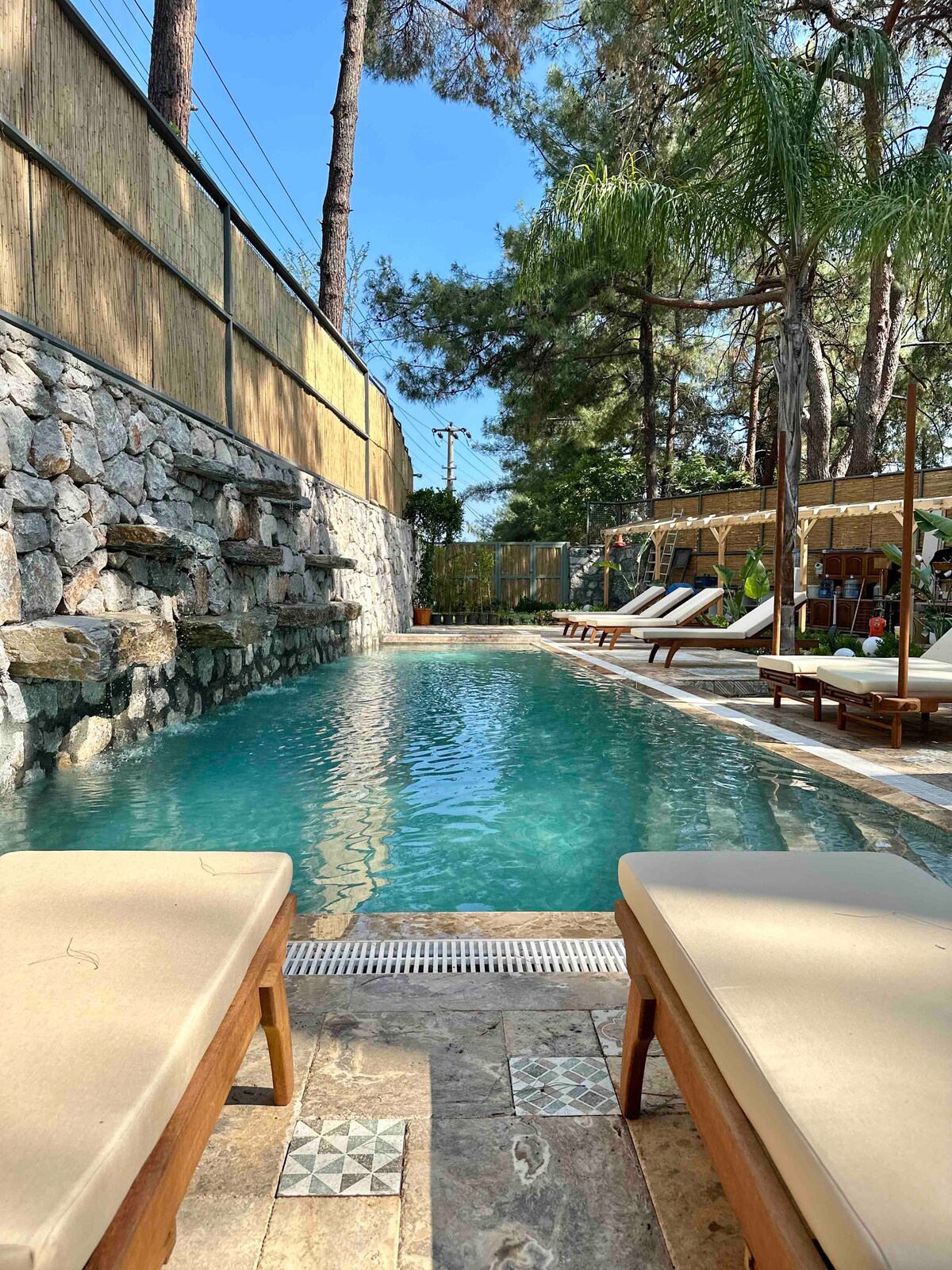 A serene outdoor pool area is surrounded by natural stone walls and vibrant greenery. Lounge chairs are positioned poolside, inviting relaxation. Sunlight reflects off the clear water, creating a calming atmosphere, while a shaded seating area with umbrellas is visible in the background.
