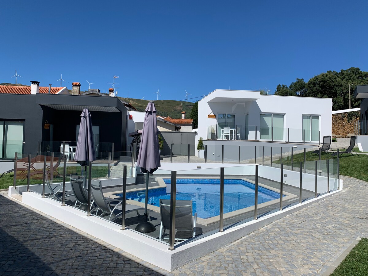 A refreshing swimming pool is surrounded by a glass barrier, featuring lounge chairs and umbrellas for shade. The modern villa with large windows stands nearby, complemented by a clear blue sky and scenic views of the hills in the background.