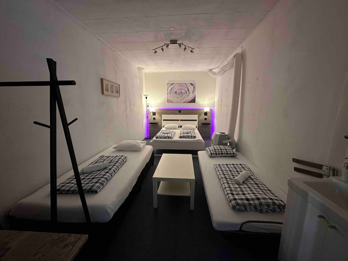 A spacious room is arranged with three beds, each adorned with checkered bedding. A minimalist table sits at the center, while soft lighting creates a calming ambiance. A wall-mounted artwork adds a touch of character, complemented by wooden accents in the furnishings.