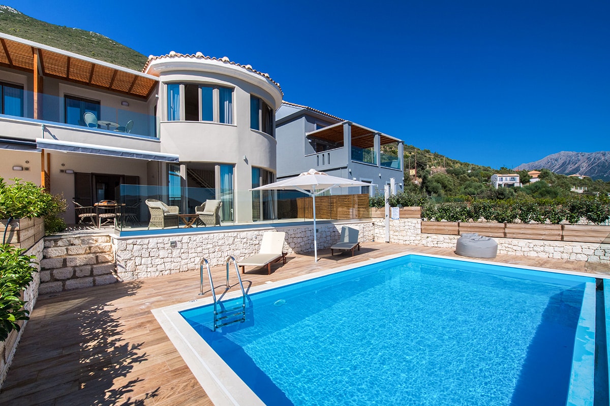 The outdoor area features a private swimming pool with clear blue water, surrounded by a wooden deck. Sun loungers are positioned nearby. The stylish villa is partially visible with large windows and a curved design, offering views of the surrounding landscape and mountains.