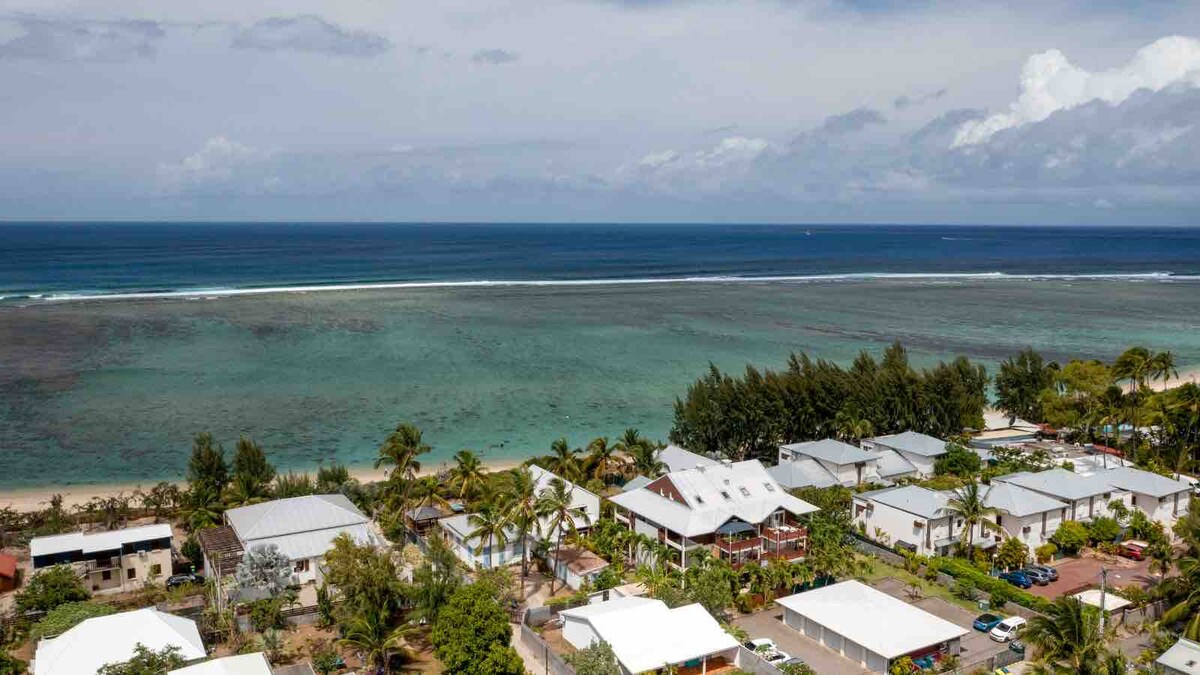A panoramic view captures the serene coastline with gently rolling waves and vibrant blue water. Lush greenery and palm trees are visible in the foreground, while a cluster of houses with white roofs is set against the backdrop of the ocean and sky.