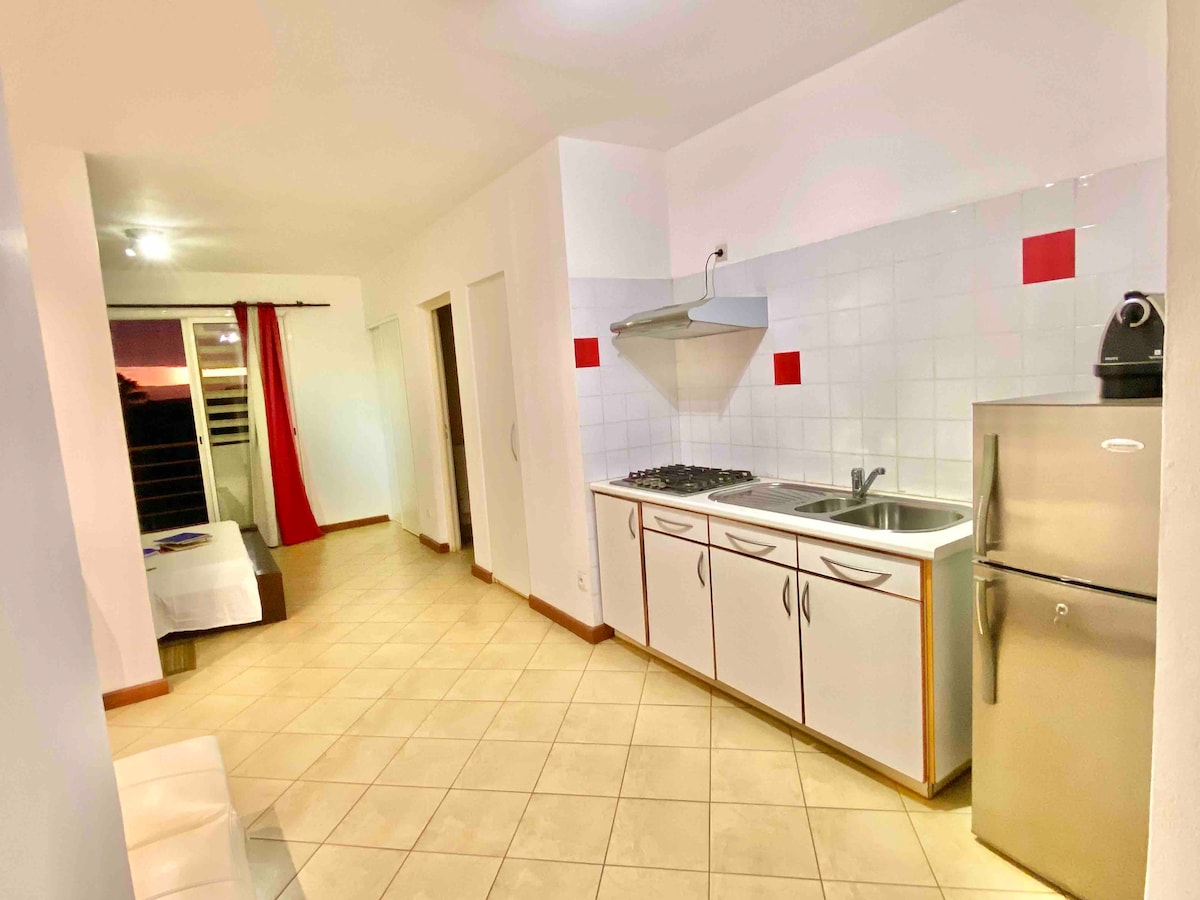 An open kitchen area features modern appliances, including a stainless-steel refrigerator and gas stove. White cabinetry is complemented by red accent tiles. A spacious living area can be seen in the background, designed with neutral tones and ample natural light from sliding doors.