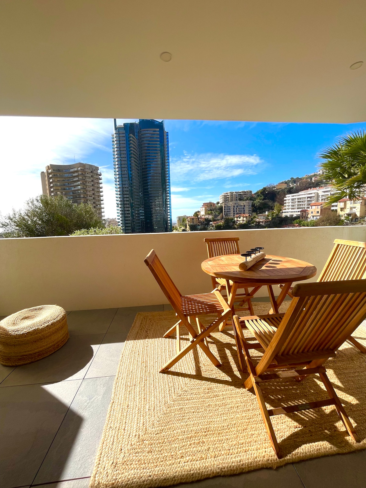 A private terrace features a round wooden table with four matching chairs, complemented by a natural fiber rug. Views of modern buildings and palm trees are framed by an expansive window, allowing natural light to fill the space.
