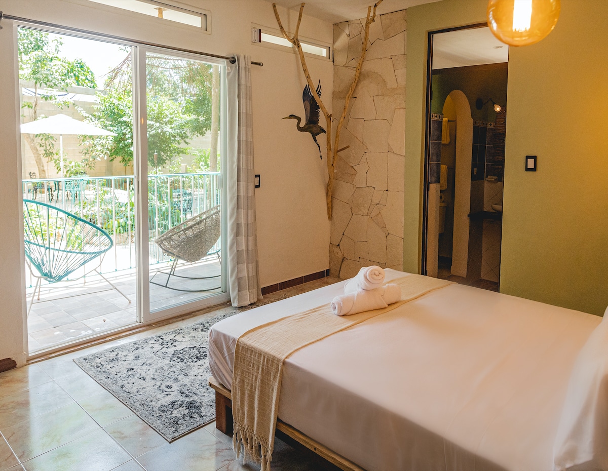 A bright and airy private room features a spacious bed covered with a light blanket, accompanied by rolled towels. Large sliding glass doors lead to a balcony overlooking a garden, while a decorative bird sculpture adds a touch of charm to the stone wall.