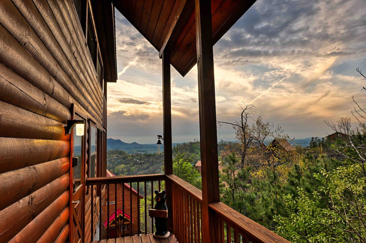 A covered porch offers views of the surrounding landscape, featuring trees and distant mountains under a sunset sky. The wooden railing provides a natural accent, with a lantern mounted to the cabin wall contributing to the warm ambiance.