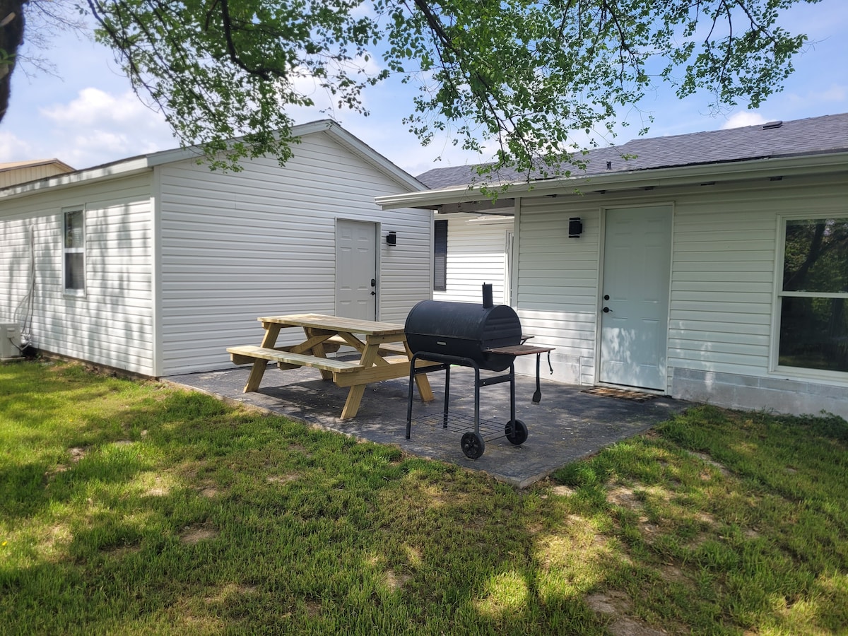 A spacious outdoor area features a picnic table adjacent to a charcoal grill. The area is bordered by green grass and is flanked by two white buildings. Sunlight filters through the leaves of nearby trees, creating a relaxed, inviting space.