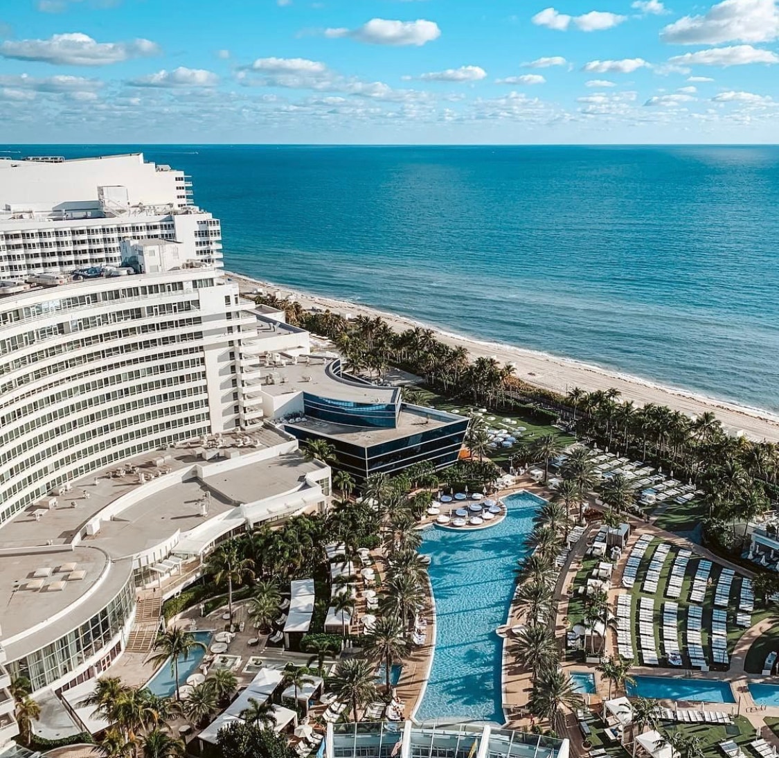 An aerial view showcases a resort complex with a large swimming pool surrounded by palm trees and lounge chairs. The ocean stretches out in the background, meeting a clear blue sky dotted with clouds. The beach is accessible beyond the pool area.