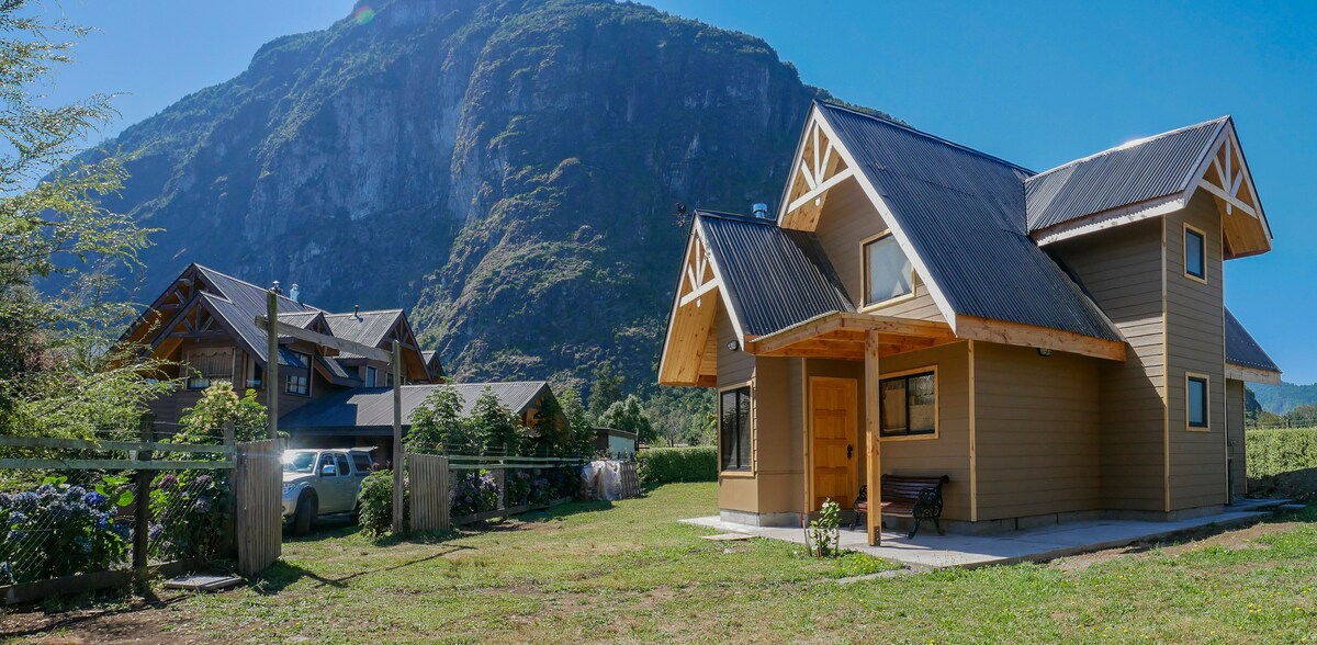 A two-story cabin is shown with a welcoming entrance and a covered porch. The structure is set against a backdrop of towering mountains, emphasizing its natural surroundings. Lush green grass and various plants enhance the outdoor space, while nearby cabins are visible in the background.