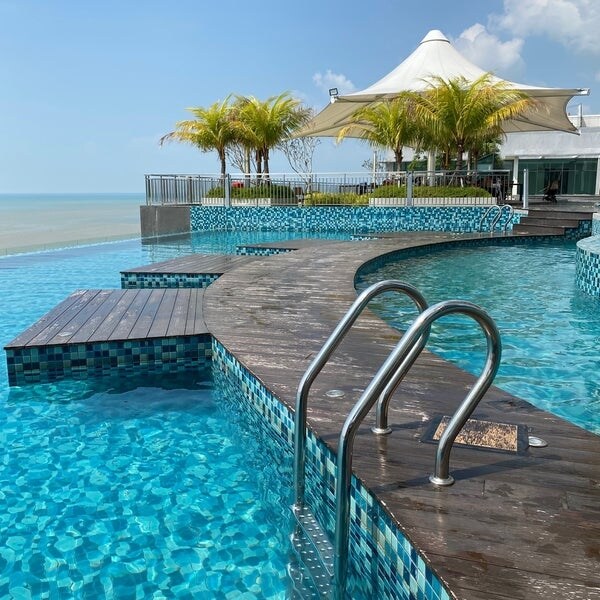 A curved swimming pool featuring a wooden pathway and two metal handrails offers access to the water. Lush palm trees are seen in the background, alongside a shaded seating area under a large white canopy, with the ocean visible in the distance.