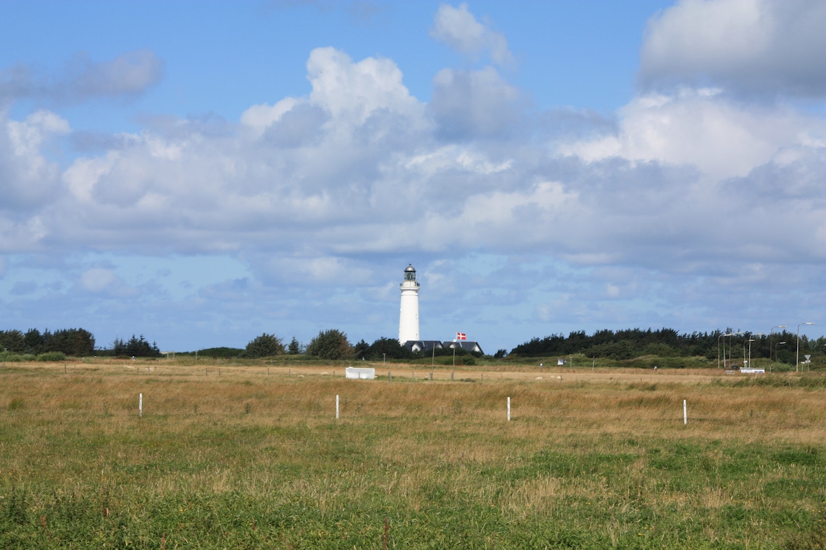 A white lighthouse rises in the distance against a backdrop of blue sky and scattered clouds. Surrounding grassland displays a mix of green and gold hues, with the gentle hills leading to the coastline. The image conveys a serene rural landscape.