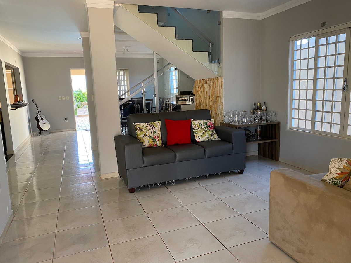 A spacious living area is presented with a gray sofa adorned with colorful cushions, positioned on glossy tile flooring. Natural light enters through a large window, illuminating a minimalist design that includes a staircase and a side table with glassware.