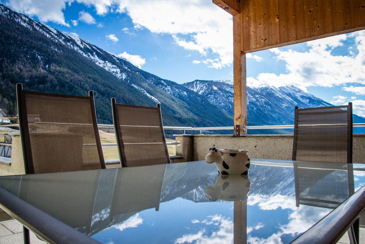 A glass table is positioned on the terrace, reflecting the surrounding mountains. Four metal chairs with mesh backs are arranged around the table. The mountainous landscape, featuring snow-capped peaks and a partly cloudy sky, creates a scenic backdrop.