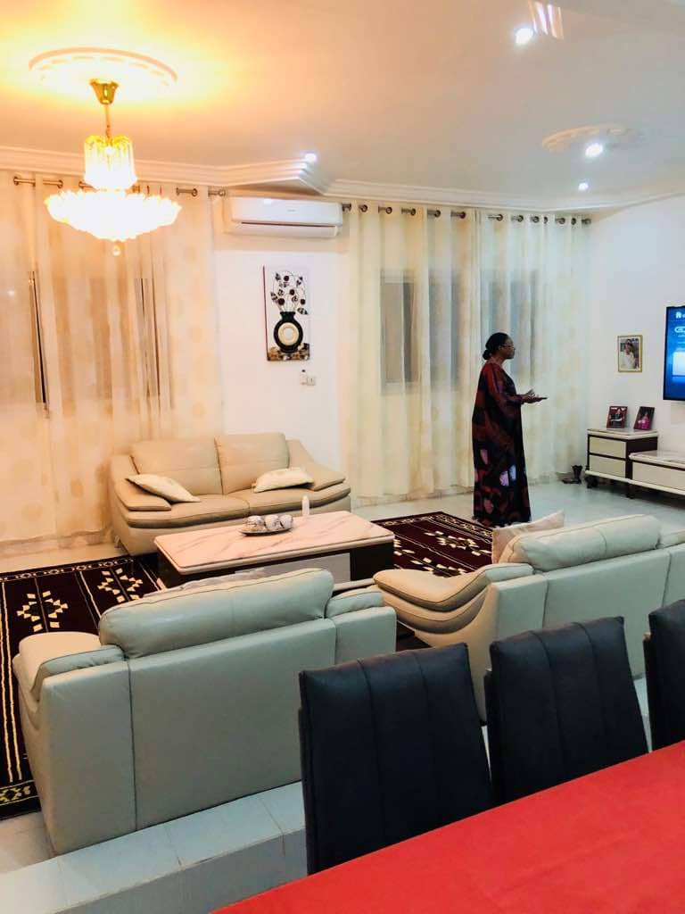 A spacious living area is furnished with light-colored sofas and a coffee table on a richly colored rug. Tall, sheer curtains allow natural light to filter in, and wall art is displayed nearby. A television is visible, enhancing the comfortable ambiance of the space.