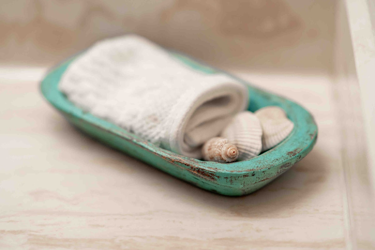 A rustic, aqua-colored dish is gently worn, holding a rolled white towel alongside an assortment of seashells. The soft textures and natural elements create a serene coastal ambiance.