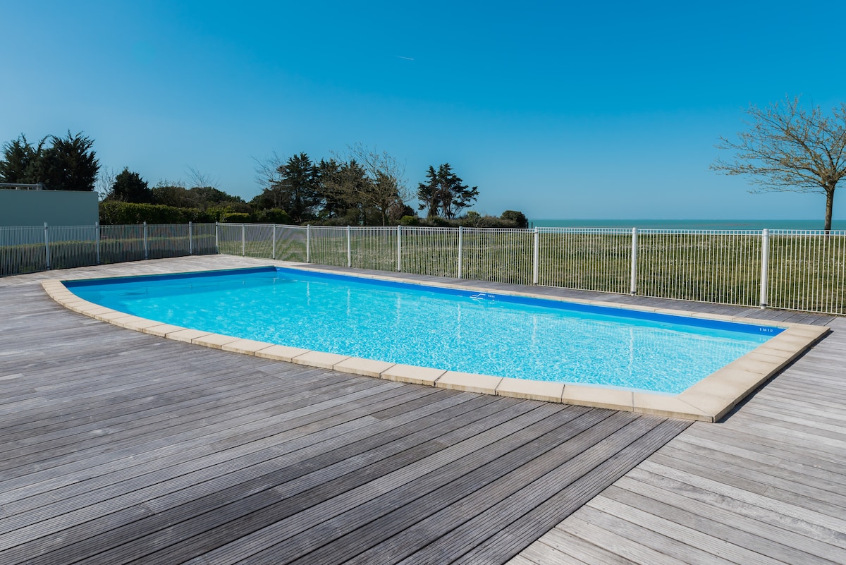 The pool is surrounded by a wooden deck, featuring a clear blue water surface reflecting the sky. A white fence provides safety while offering unobstructed views of the nearby green landscape and ocean in the background.