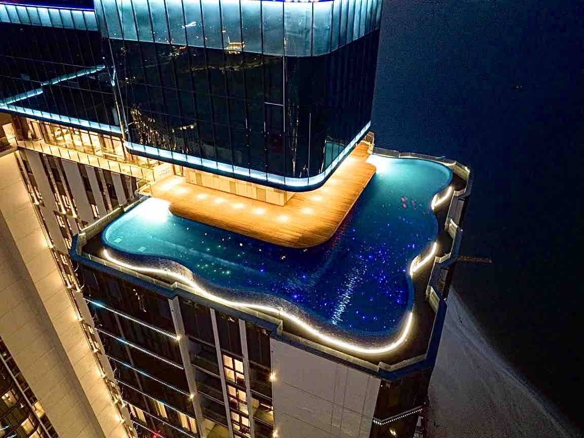 An expansive rooftop infinity pool is illuminated at night, surrounded by glowing lights that enhance the architectural features of the building. The pool's water reflects the city lights, creating a serene atmosphere above the bustling surroundings.