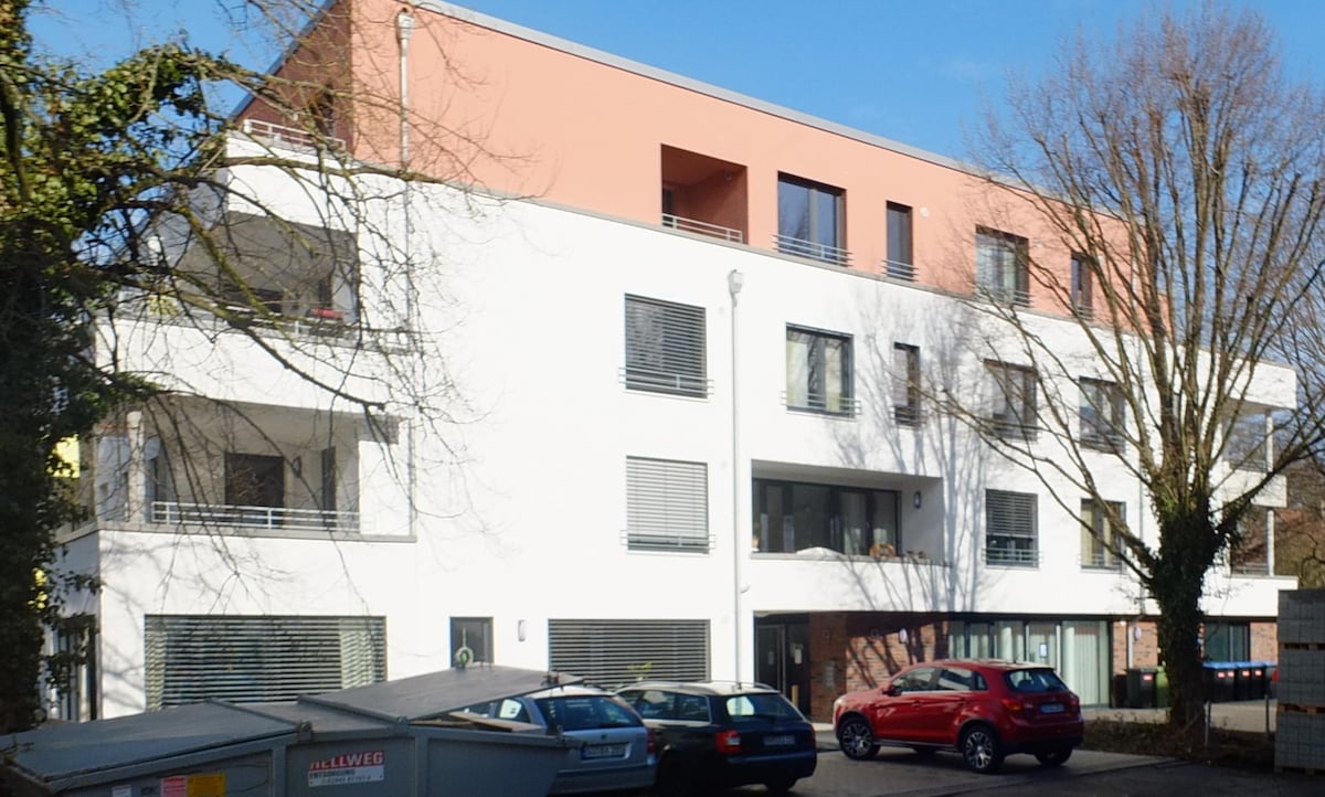 The modern multi-story building showcases a combination of white and orange façades, framed by large windows. Surrounding greenery enhances the environment. The parking lot is visible in the foreground, featuring vehicles parked along the side of the structure.