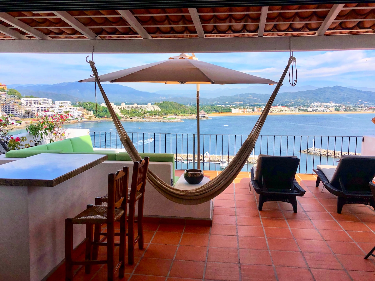A spacious patio features a hammock beneath a large umbrella, offering views of the coastline and mountains. Outdoor seating includes a bar and lounge chairs, with vibrant green cushions. The tiled flooring complements the relaxing atmosphere, inviting guests to enjoy the surroundings.