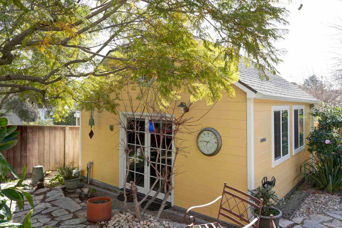 The exterior of a cheerful yellow cottage is framed by lush greenery and trees. A stone pathway leads to the entrance, which features a large clock on the wall and a decorative chair in the foreground, inviting a serene outdoor atmosphere.