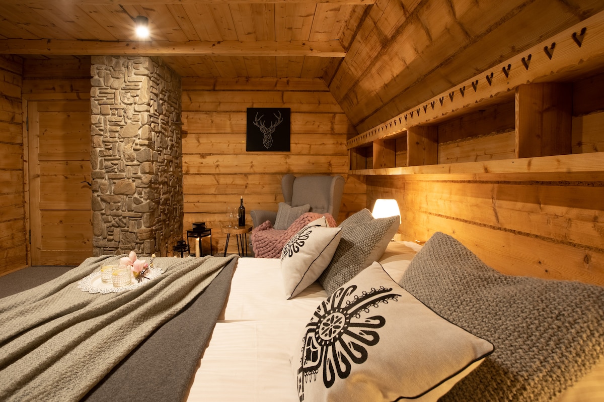 A warm wooden interior features a spacious bed adorned with decorative pillows and a textured blanket. Soft lighting highlights the stone accent wall and built-in shelving, creating a cozy atmosphere. A small table sits nearby, complemented by a bottle and glasses.