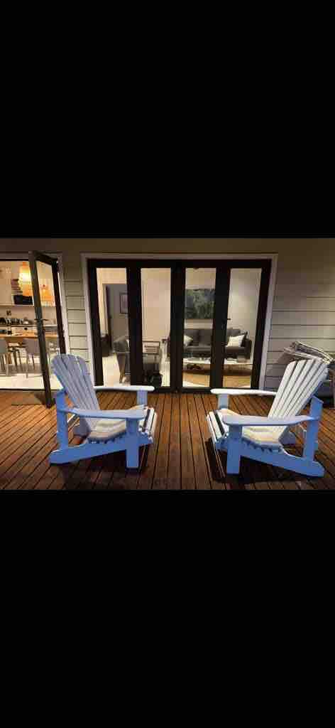 Two light blue Adirondack chairs are positioned on a wooden deck, facing a set of glass doors that reveal a living area inside. The warm light from the interior illuminates the inviting space, creating a connection between indoor and outdoor relaxation areas.