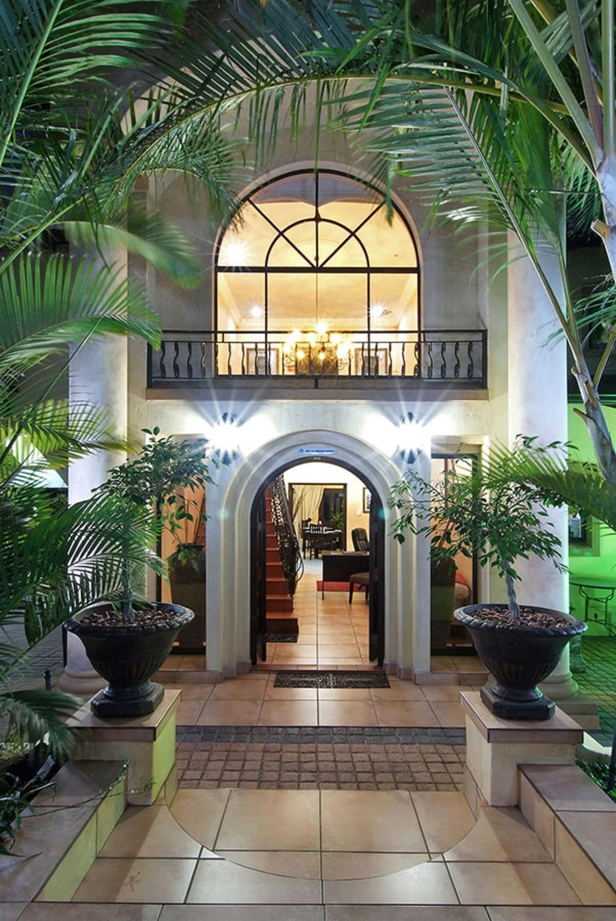 The entrance showcases an elegant archway framed by lush green palms. The tiled pathway leads to a spacious foyer visible through large windows above, where indoor lighting creates a welcoming ambiance. Stone planters hold potted plants, enhancing the inviting atmosphere.