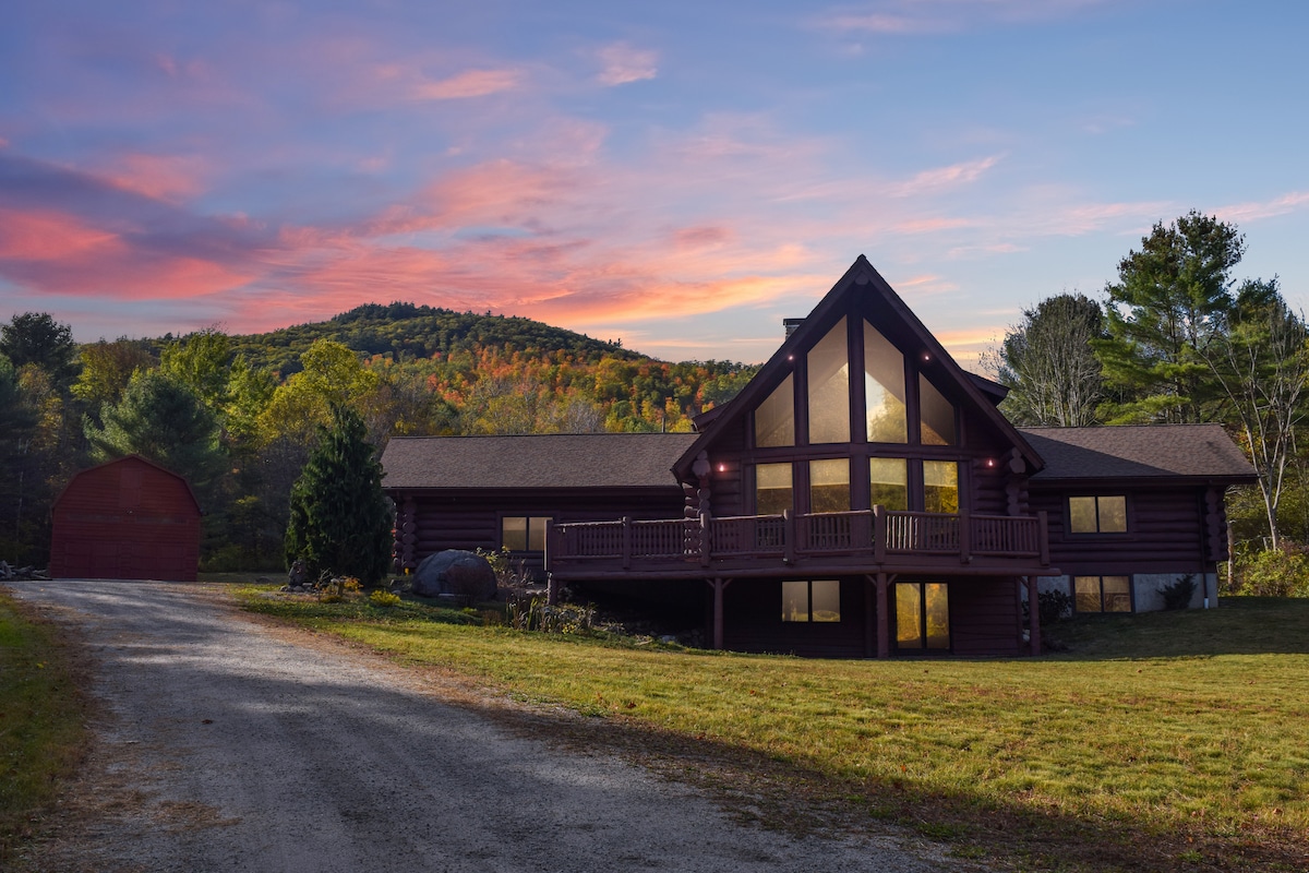 A log cabin chalet is depicted, featuring large windows that reflect the surrounding landscape. The home is set on a spacious lawn, leading to a gravel driveway that connects to a barn. Soft colors of sunset illuminate the mountain backdrop.